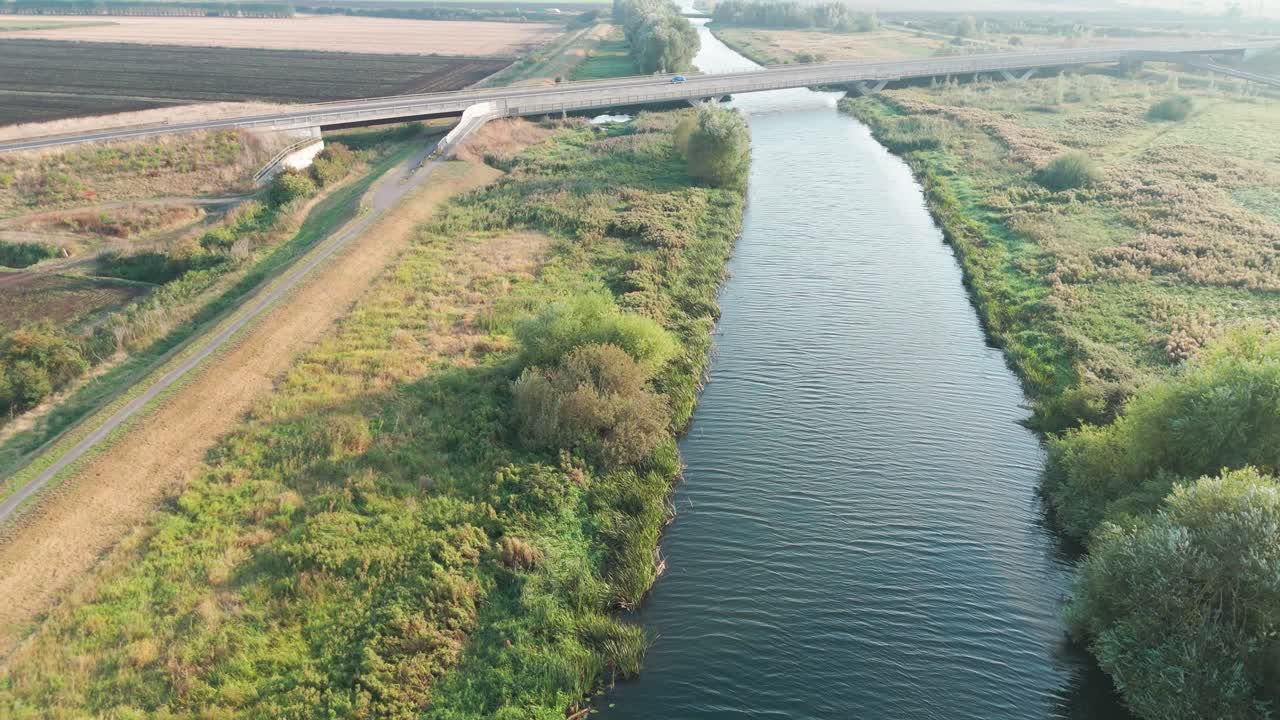 Aerial view of a serene river flowing under a bridge with adjacent green fields and a country road. The landscape features trees, bushes, and grass, creating a tranquil rural scene under a blue sky