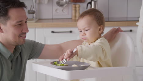 Padre-Alimentando-A-Su-Niña-Feliz-Sentada-En-Su-Silla-Alta-En-La-Cocina