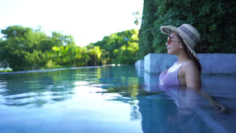 Beautiful-young-Asian-woman-sitting-in-shaded-swimming-pool-wearing-light-colored-bikini,-sunglasses-and-straw-hat-looks-out-on-tropical-landscape-of-a-luxury-resort-in-an-island-paradise