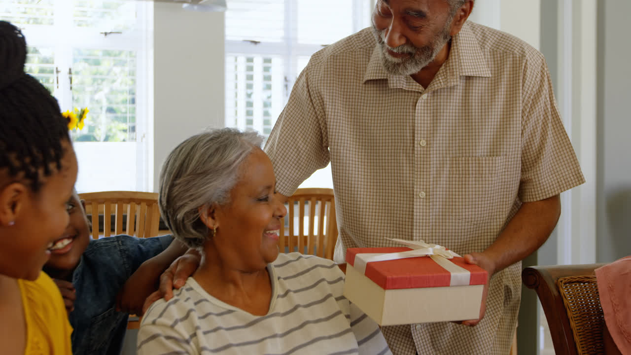 Front view of senior black man giving gift to senior woman at dining table in a comfortable home 4k