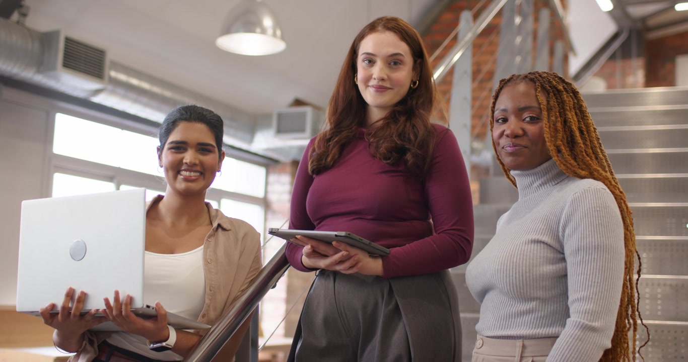 Squadra femminile eterogenea in piedi sulle scale, con la donna al centro che tocca il tablet e conduce una revisione con il laptop