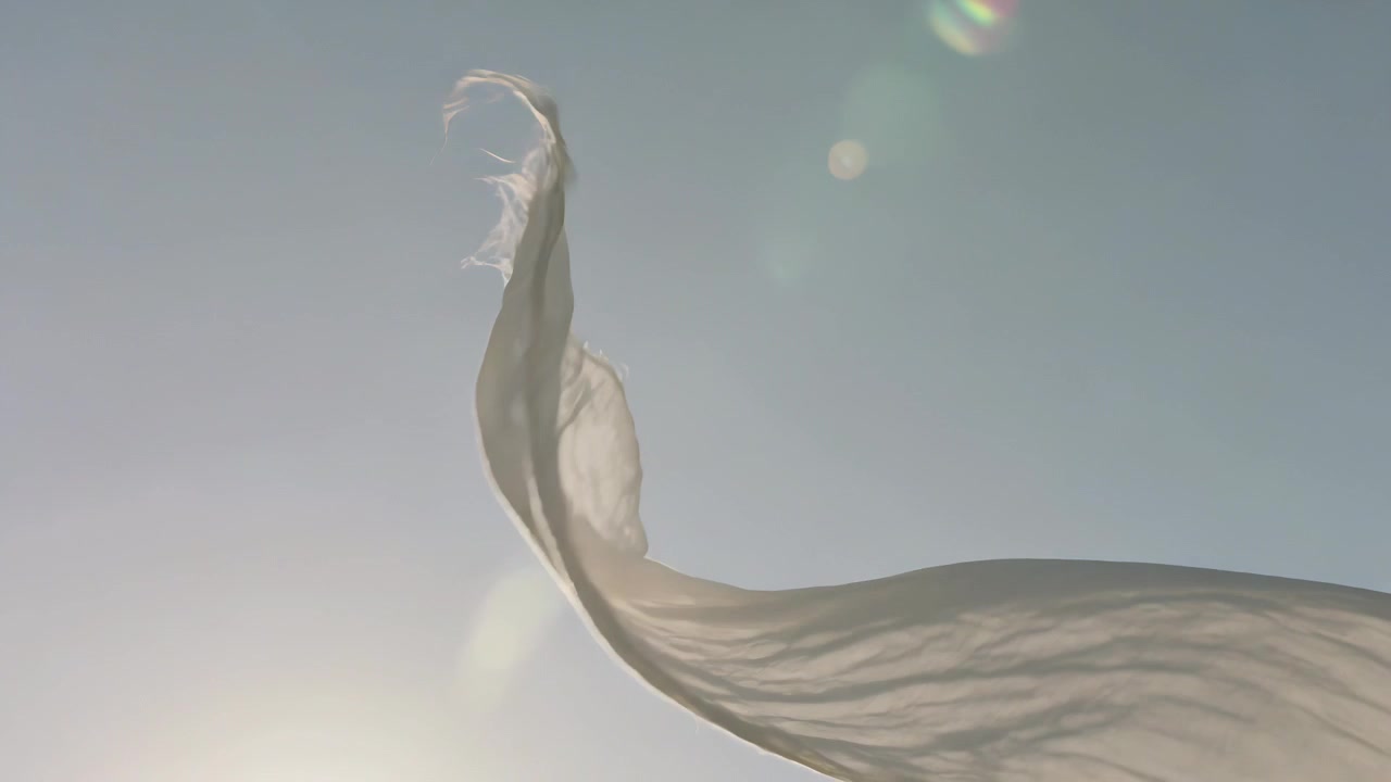 Weißes Laken weht im Sonnenlicht und Windhauch zum Himmel, zeigt ausgefranste Kanten und Linsenreflexionen.