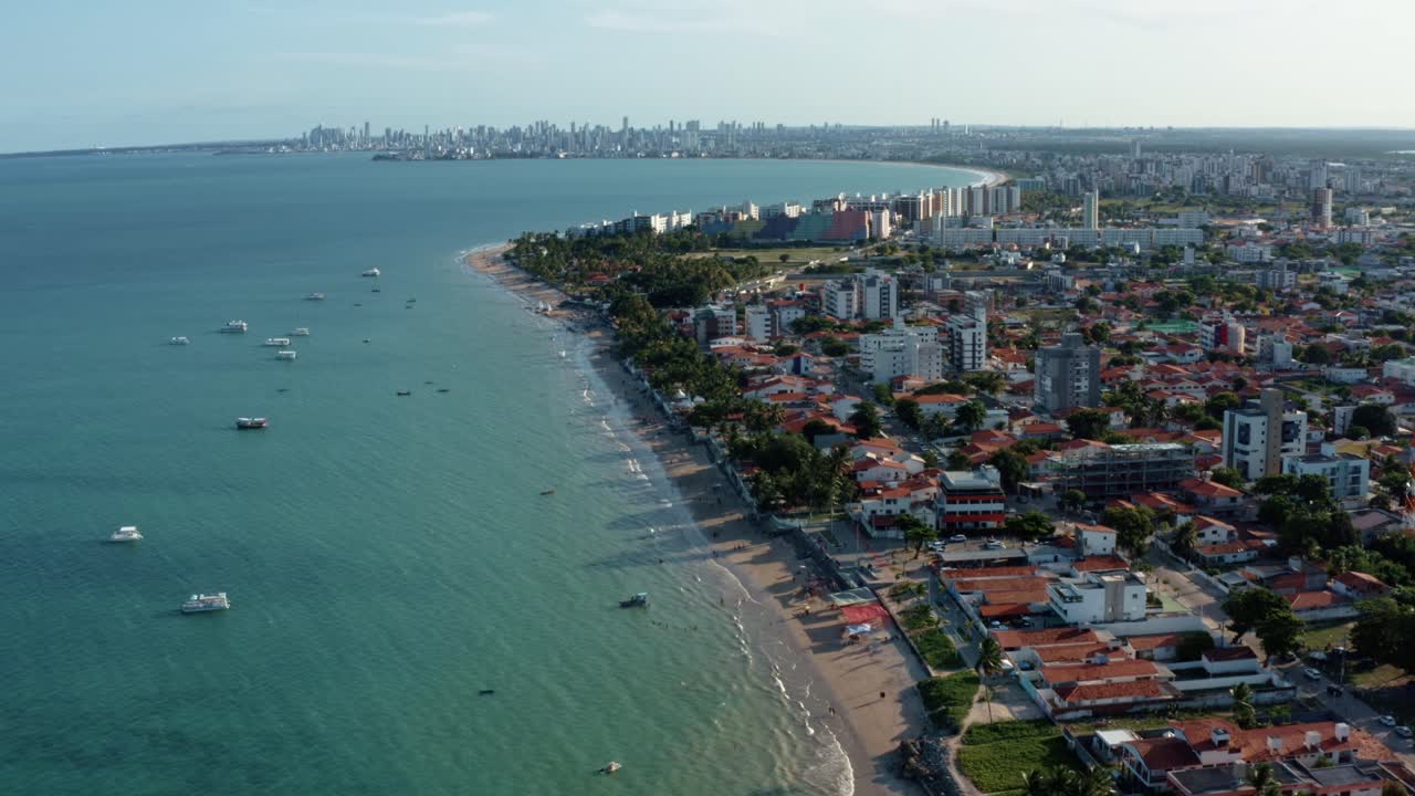 Tilt up aerial drone extreme wide shot of the tropical Bessa beach in the city of Joao Pessoa, Paraiba, Brazil with people enjoying the ocean, tour boats, and large cityscape in the background