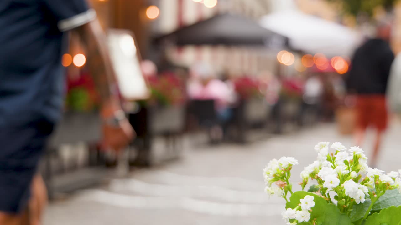 People walk past white blossoms and outdoor tables on a lively, softly lit cobblestone street