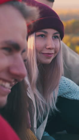 positive blonde girl spends time with boyfriend and mates at burning bonfire on meadow in warm autumn evening closeup