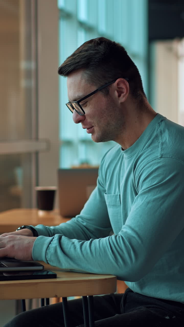Freelancing man working on his laptop sitting in a cafe. Male in glasses typing on computer in a relaxed atmosphere. Blurred backdrop. Vertical video
