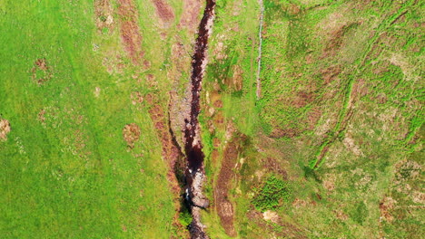 Toma-Aérea-De-Arriba-Hacia-Abajo-Siguiendo-Un-Arroyo-Rocoso-En-Medio-Del-Campo-Verde,-Día-Soleado-Y-Brillante