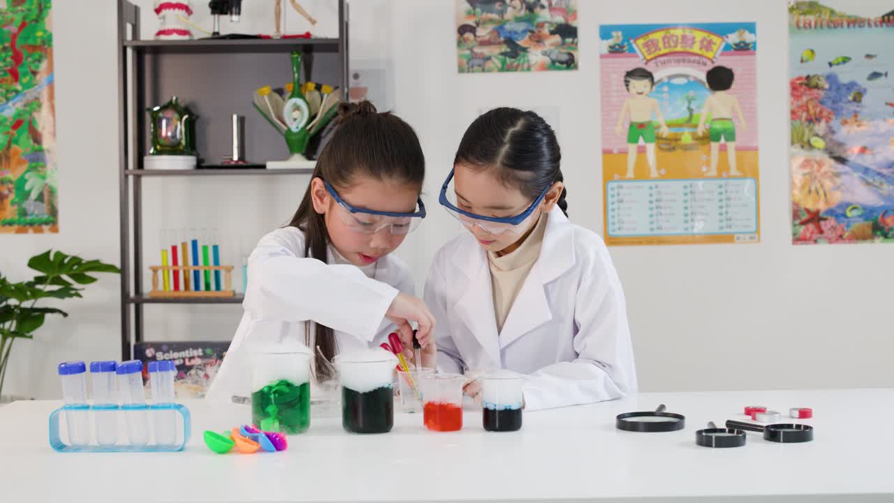 Dua gadis muda Asia mengenakan jas lab dan kacamata pelindung saat menggunakan pipet di dalam kelas