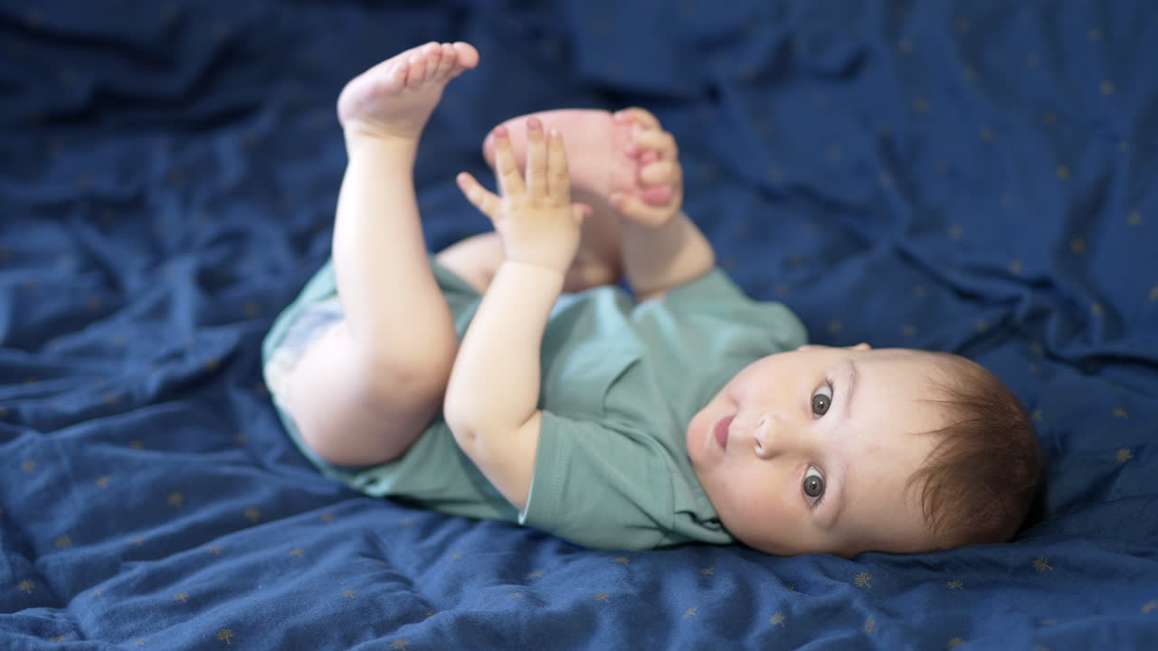 Caucasian baby boy lies on the bed holding legs up. Active child playing with his feet and toes. Blue backdrop.