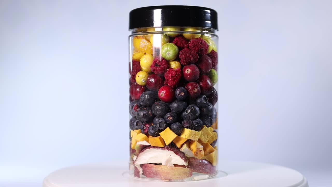 A transparent jar filled with a colorful assortment of freeze-dried fruits and vegetables, including blueberries, cranberries, raspberries, apple slices, pumpkin cubes, and more.