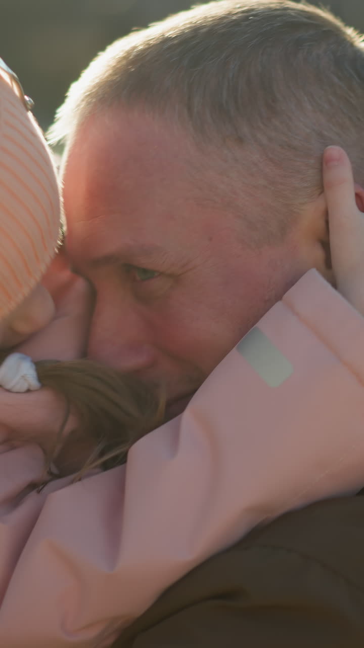 un momento conmovedor capturado cuando una niña con una gorra y chaqueta rosas coloca sus manos amorosamente en la cara de un hombre mientras es llevada. la luz del sol agrega un caloroso resplandor a esta interacción sincera