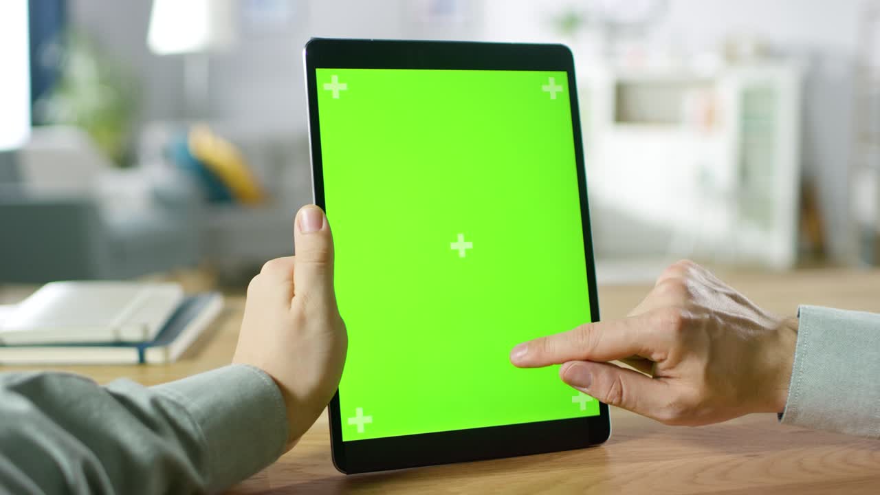 primer plano de un hombre usando gestos con las manos en una pantalla de maqueta verde, una tableta digital en modo retrato mientras está sentado en su escritorio. en el fondo, una acogedora sala de estar.