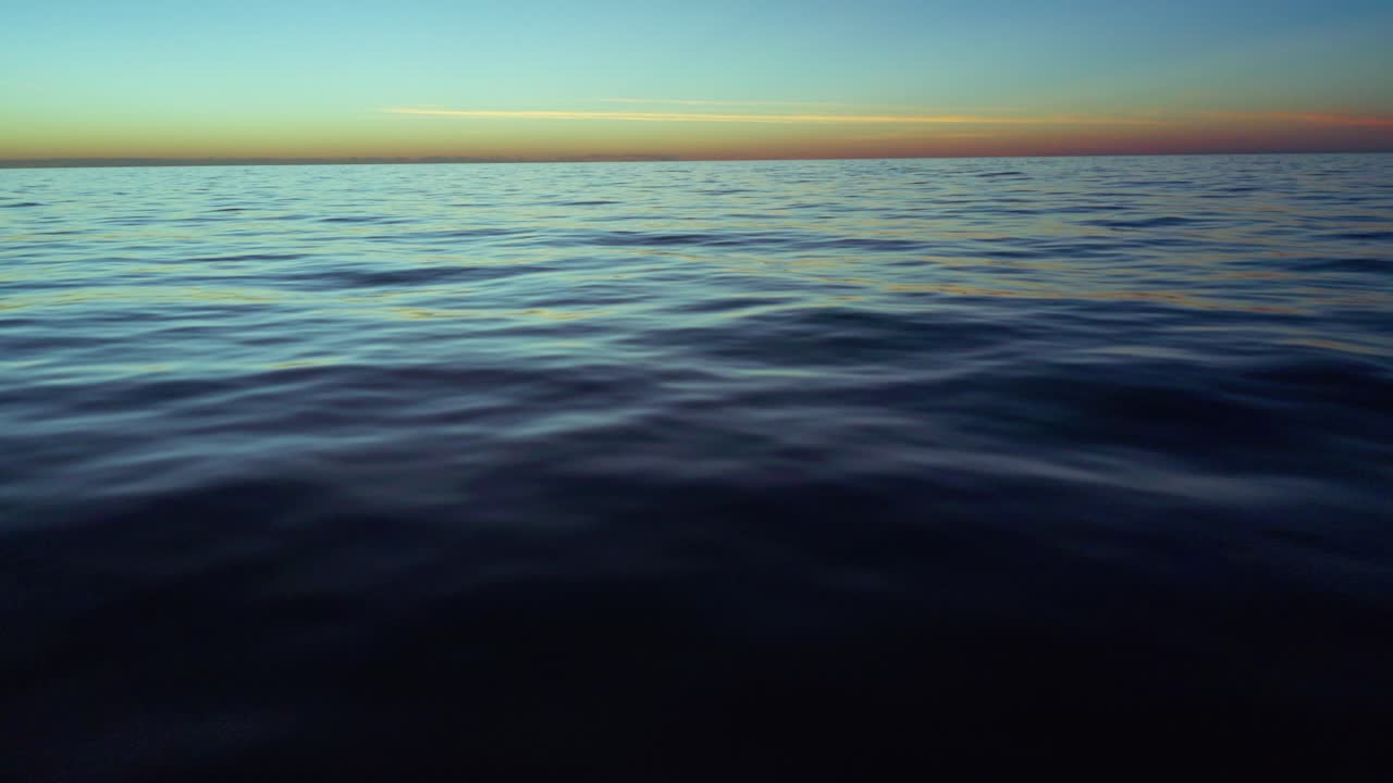 Ocean surface reflects soft light as the sun transitions during sunset or sunrise, with gentle waves and a wide horizon under a colorful sky