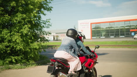 a woman in a checkered shirt rides a red bike with her friend holding onto her from behind, both wear helmets, with trees by the side, and a distant building with glass