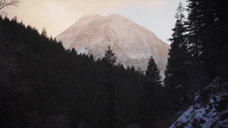 Tilt-up-revealing-a-gorgeous-vivid-fire-like-sunset-with-Mount-Timpanogos-glowing-and-large-pine-trees-surrounding-on-a-cold-snowy-winter-evening-up-American-Fork-Canyon,-Utah