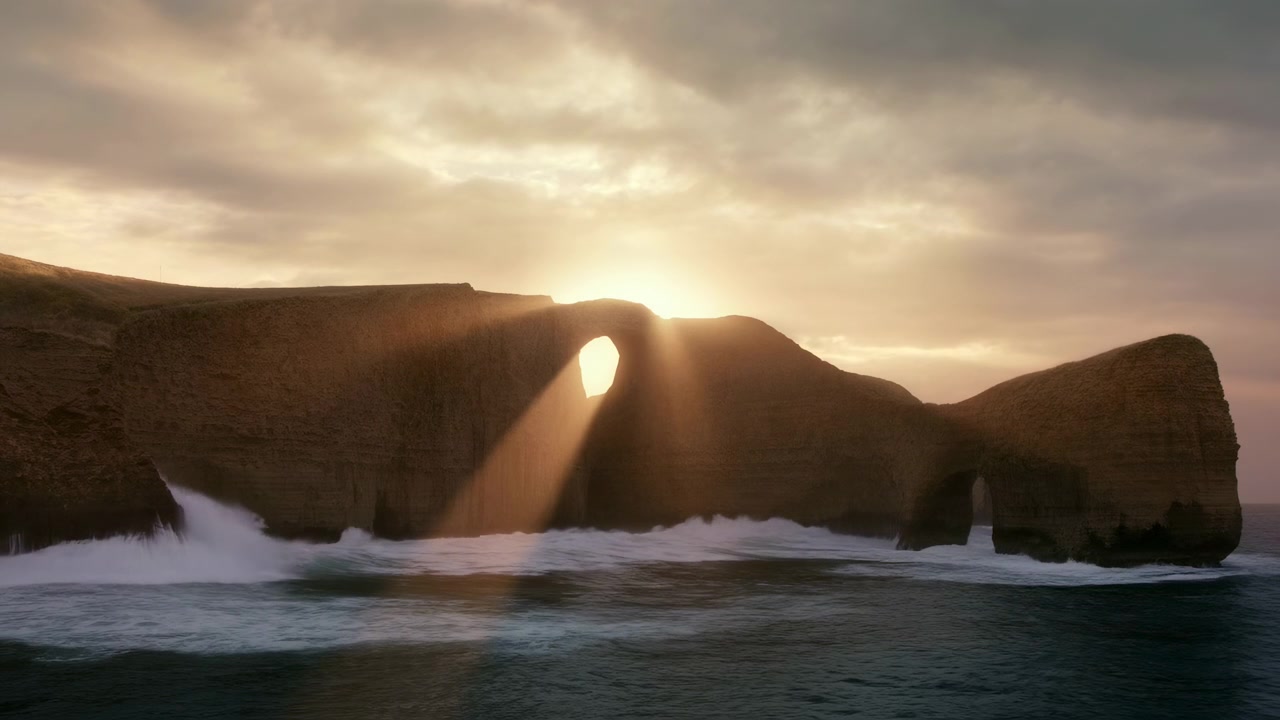 Scena costiera panoramica ripresa dal basso con una grande scogliera sedimentaria e un arco di roccia circolare vicino al centro, mentre il sole si trova dietro l'apertura, proiettando una luce dorata attraverso l'arco mentre le onde si infrangono e gli spruzzi si disperdono.