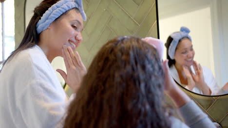 Happy-biracial-mother-and-daughter-with-headbands-putting-cream-on-faces-in-sunny-bathroom