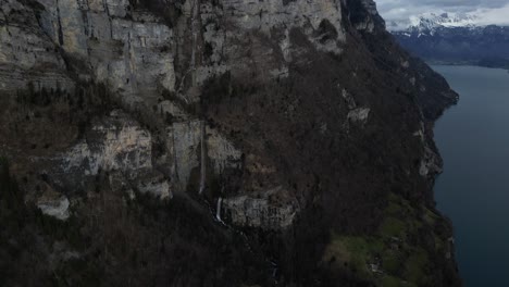 Drone-shot-of-cliff-of-a-hill-with-snow-covered-mountains-at-background-in-Walensee,-Switzerland