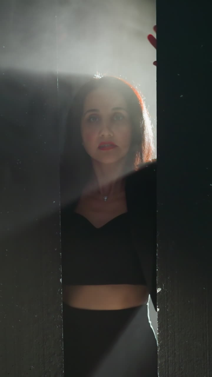 una mujer misteriosa con un traje negro emerge de las sombras, sus manos presionadas contra una pared con una luz dramática iluminando su cara mientras la cámara hace zoom.