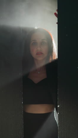 mysterious woman in a black outfit emerges from the shadows, her hands pressed against a wall with a dramatic light illuminating her face as the camera zooms in