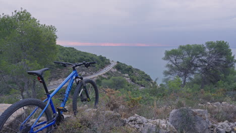 Lapso-De-Tiempo-De-La-Bicicleta-De-Montaña-Estacionada-En-La-Naturaleza-Mientras-Los-Autos-Conducen-Por-Un-Camino-Curvo-Mientras-El-Sol-Sale-Sobre-El-Océano-En-El-Fondo