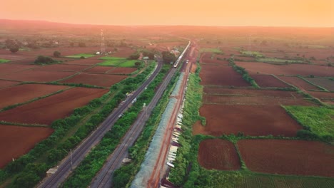 Toma-Aérea-De-Un-Dron-De-Un-Tren-De-Pasajeros-De-Ferrocarriles-Indios-Que-Pasa-Bajo-Un-Puente-De-Carretera-Durante-La-Puesta-De-Sol-En-Gwalior,-India