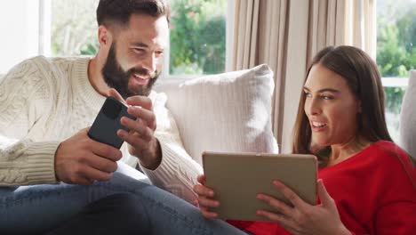 Video-of-happy-caucasian-couple-siting-on-sofa-and-using-tablet-and-smartphone