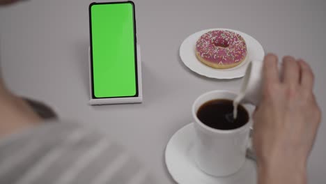 woman sitting at table coffee donut using smartphone with chroma key green screen, scrolling through social network media online shop internet. smartphone in horizontal mode with green screen mock-up.