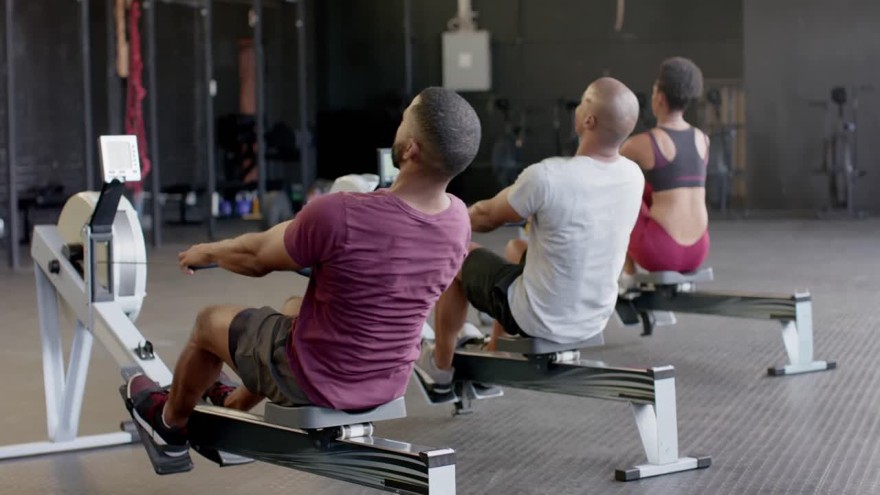 vista trasera de diversas clases de acondicionamiento físico en grupo entrenando duro en máquinas de remo en el gimnasio, en cámara lenta