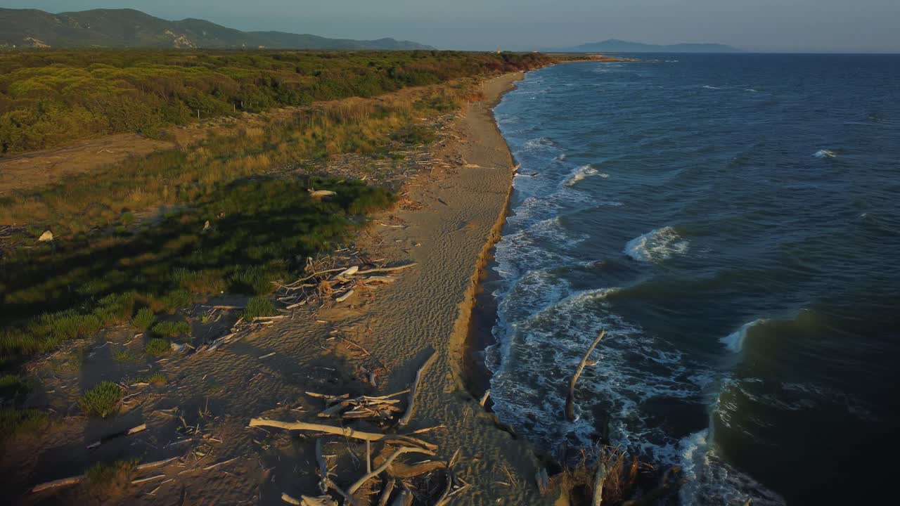Cinematic aerial drone footage flying forwards above a sandy sunrise beach at the seaside near Alberese in the iconic Maremma nature park in Tuscany, Italy, with waves, islands and a dramatic red sky