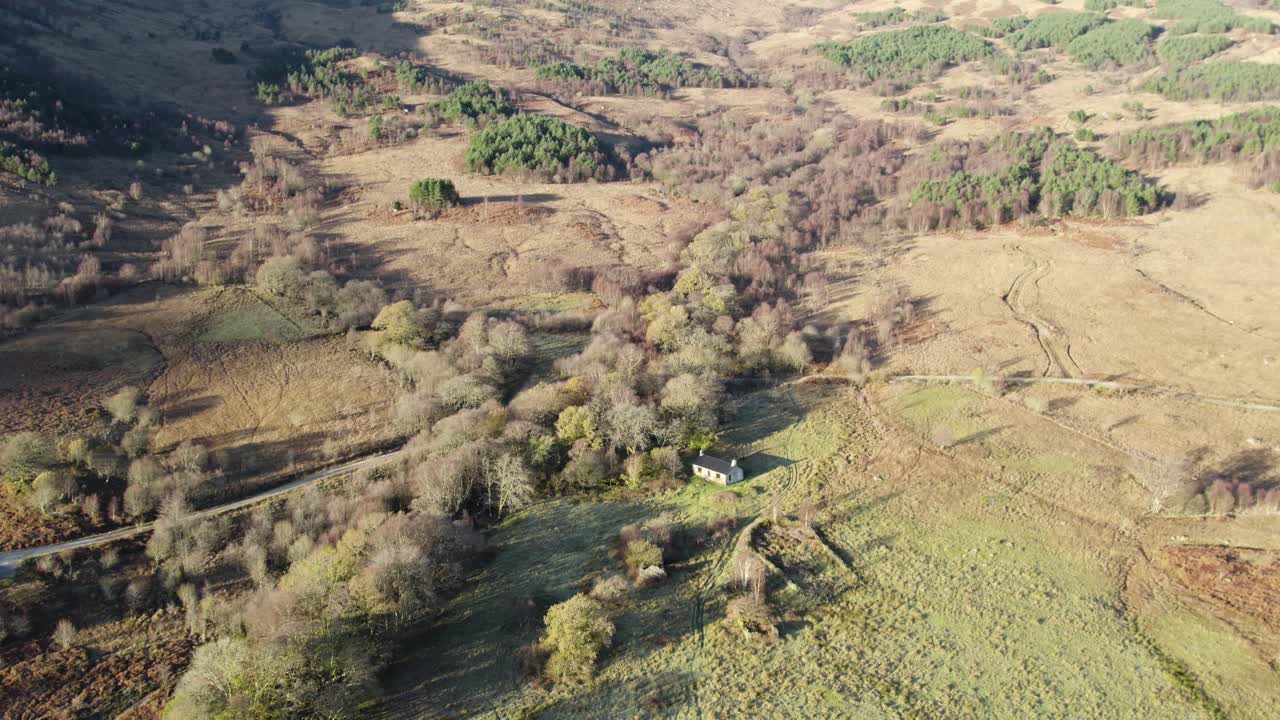 Slow aerial drone footage descending towards native woodland in winter focusing on a remote Scottish Bothy , fields, long shadows and a forested hillside in the sunshine