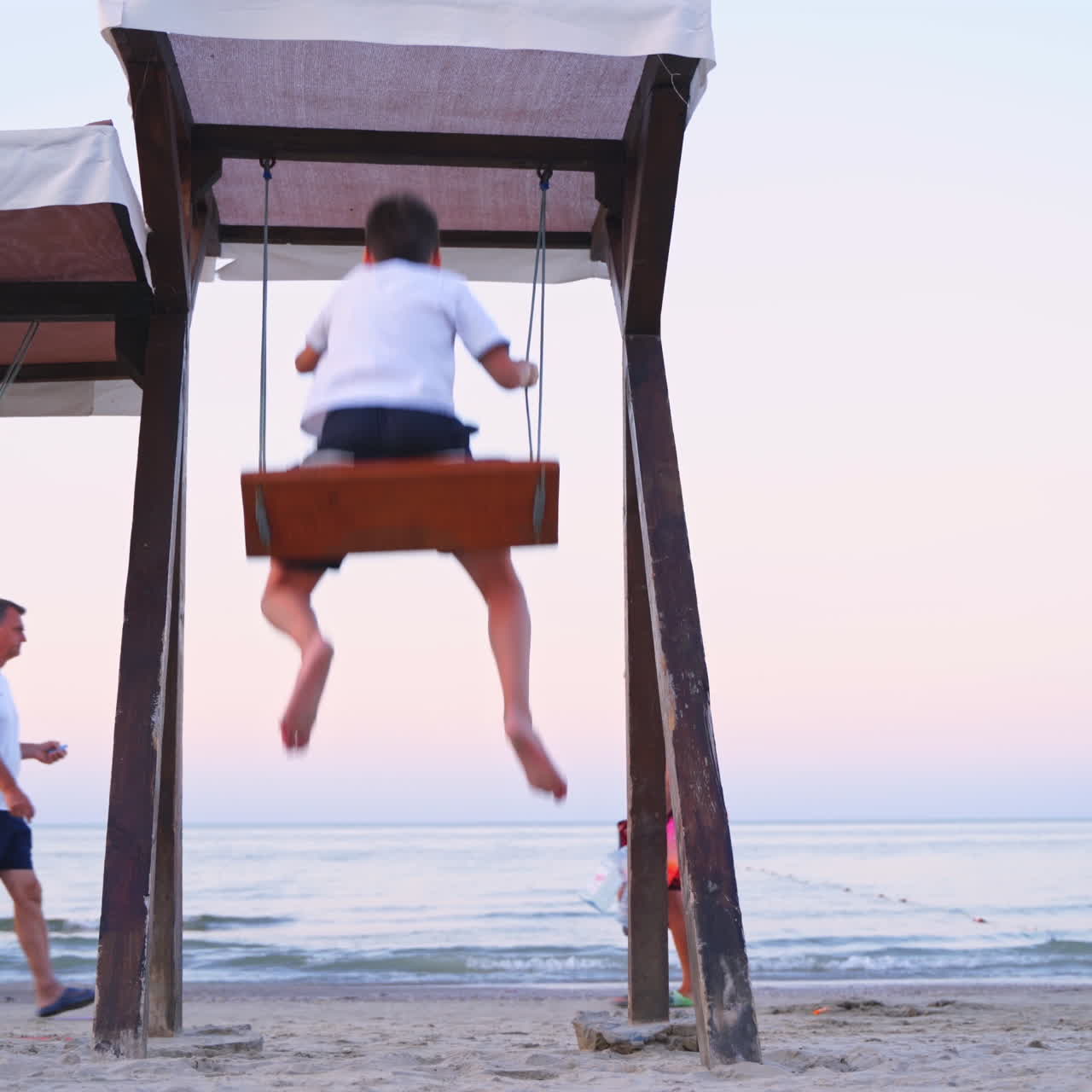 Children on a swing against the sea. Back view of a boy and girl are swinging on a playground on beach in the evening in summer.