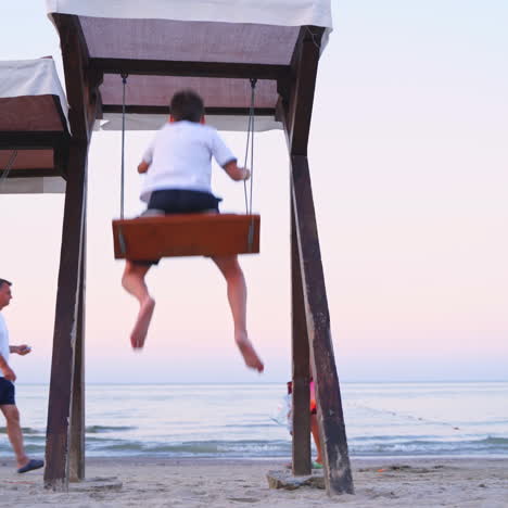 children on a swing against the sea. back view of a boy and girl are swinging on a playground on beach in the evening in summer.