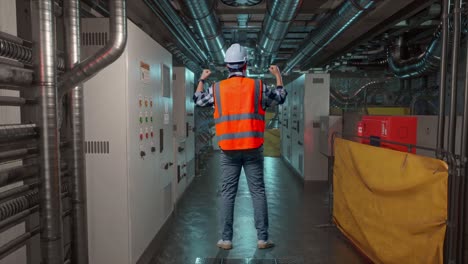 vollkörperliche rückwand eines männlichen ingenieurs mit sicherheitshelm, der seine hände erhebt, während er im motorkontrollraum arbeitet, arbeit der elektrischen generatoren