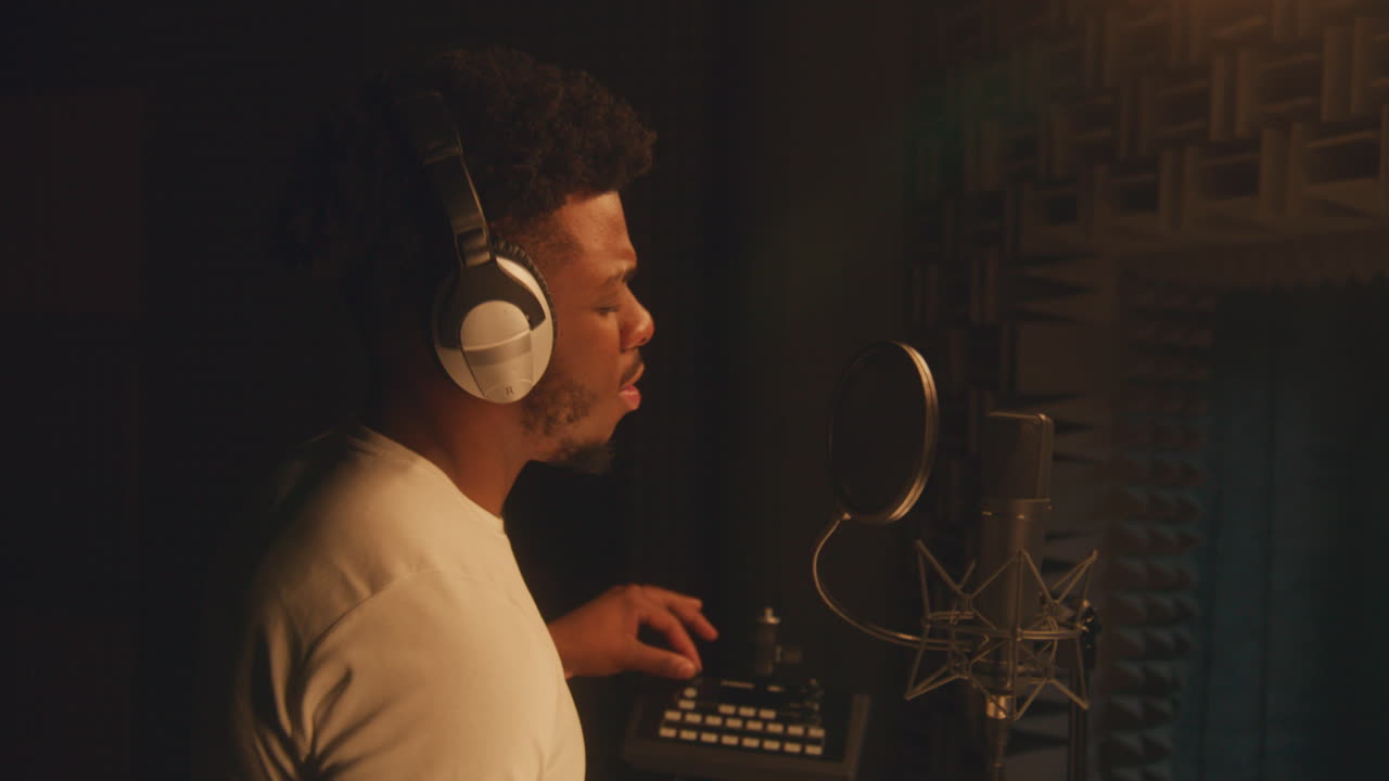 African American vocalist sings lyrical composition into microphone in soundproof room, uses mixing board and gesticulates to audio engineer. Singer works in sound recording studio. Music production.