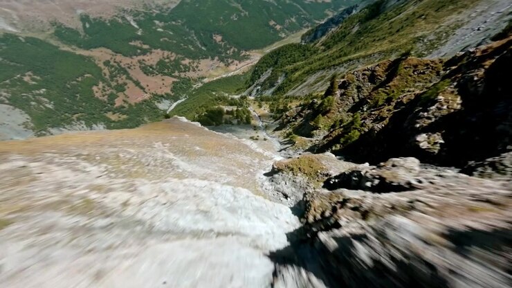 Un agile volo FPV vira oltre l'affilata cresta di una vetta e si tuffa in un'ampia valle alpina tra foreste e pendii di ghiaione