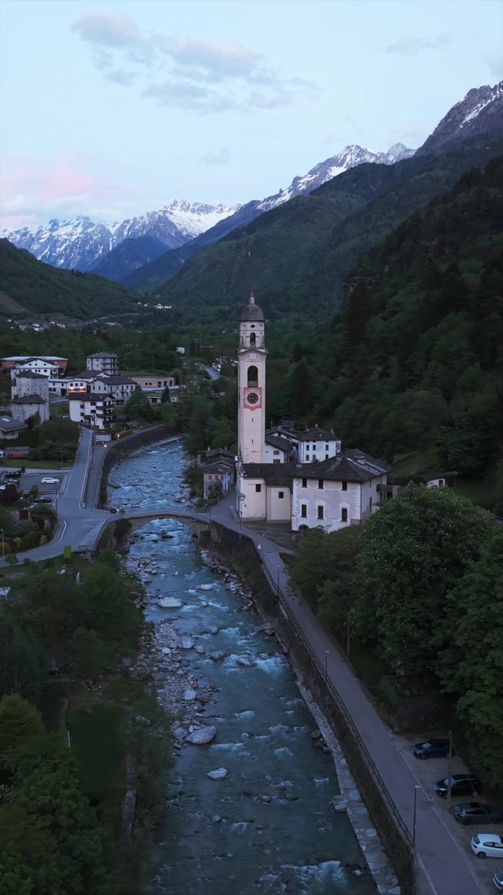 Sakin bir akşamda bir alp vadisinde, bir kilise ve köprünün yakınından akan dağ nehri, havadan dikey video.