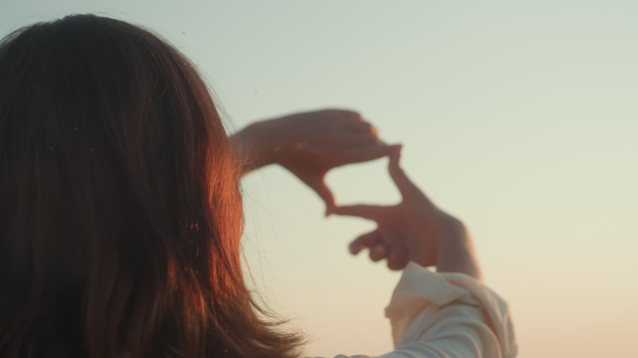 Close up rear view of woman making framing gesture toward sun, glowing rays streaming through fingers, blurred river valley and grass hill backdrop bathed in warm golden sunset light