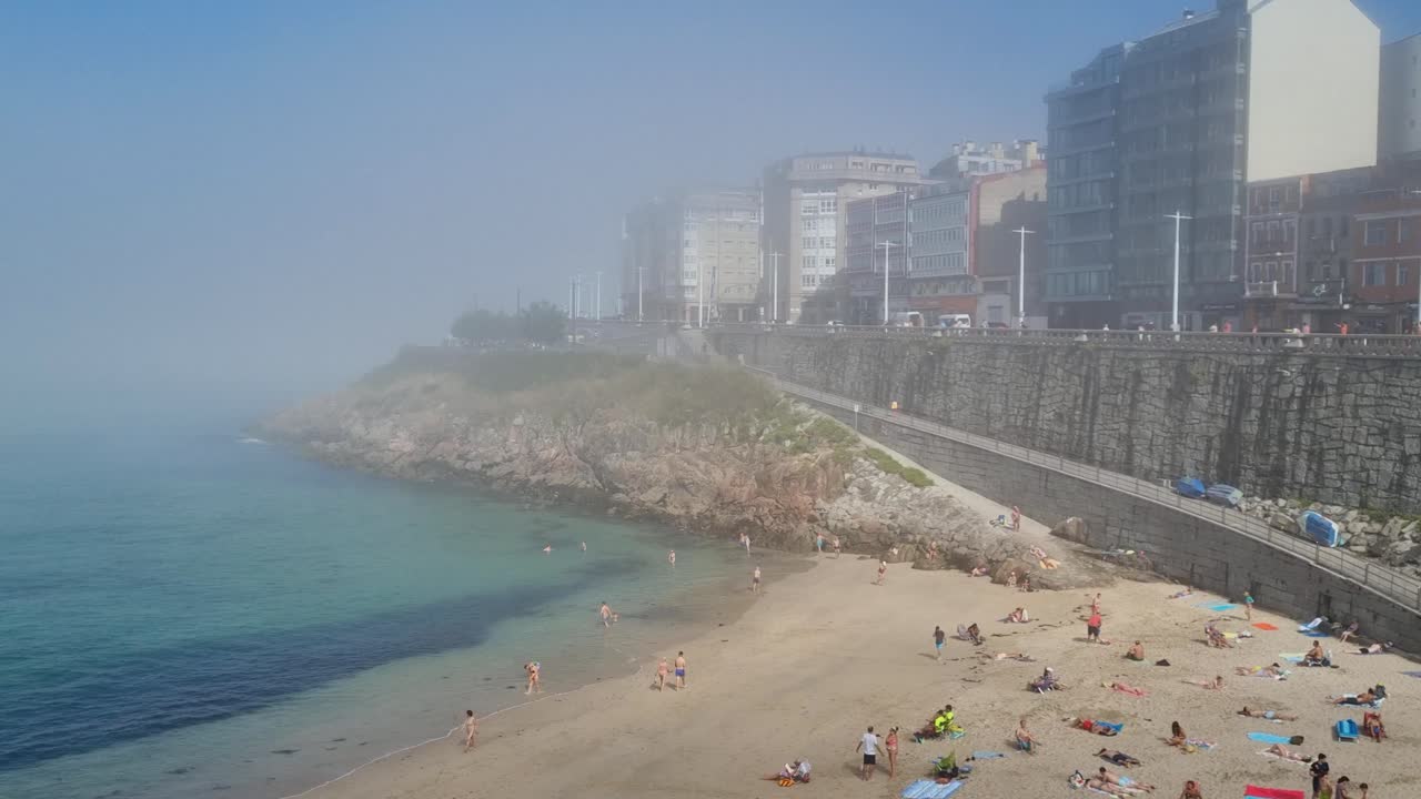 Revealing shot of beach, Atlantic sea, people sunbathing and buildings covered in mystical fog in central A Coruna Spain