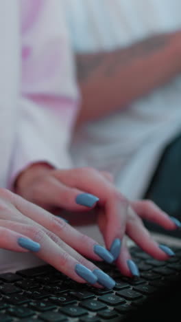 primer plano de una mujer con una camiseta rosa con uñas pulidas y vaqueros escribiendo en una computadora portátil mientras muestra algo en una tableta a un colega sentado a su lado que escribe rápidamente