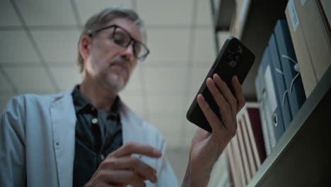hombre en bata de laboratorio usando un teléfono inteligente en una biblioteca