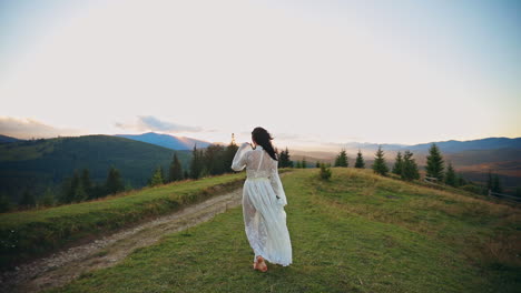 Barefoot lady in transparent white dress walks in the hills. Long-haired woman holding white flowers in her hand. Mountainous scenery with fir-trees at backdrop.