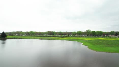 Aerial-Drone-Shot-of-Lake-Flying-Towards-Highway-and-Residential-Community-with-a-Meadow-in-Between-With-Yellow-Spring-Flowers-Starting-to-Bloom