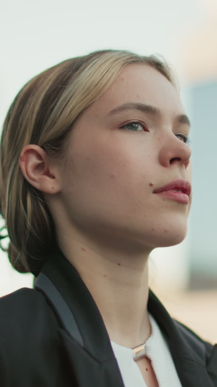 Female stylist in black suit walking with confident expression and hair clipped back, looking ahead with blurred modern cityscape in background during bright workday morning in urban environment