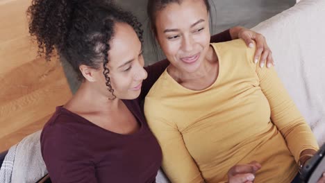 Happy-biracial-lesbian-couple-relaxing-on-couch-together,-using-tablet-and-embracing,-in-slow-motion