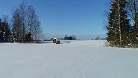 Drone-flight-over-secluded-cabin-and-thermowood-barrel-sauna-in-woods,-snowy-winter-landscape