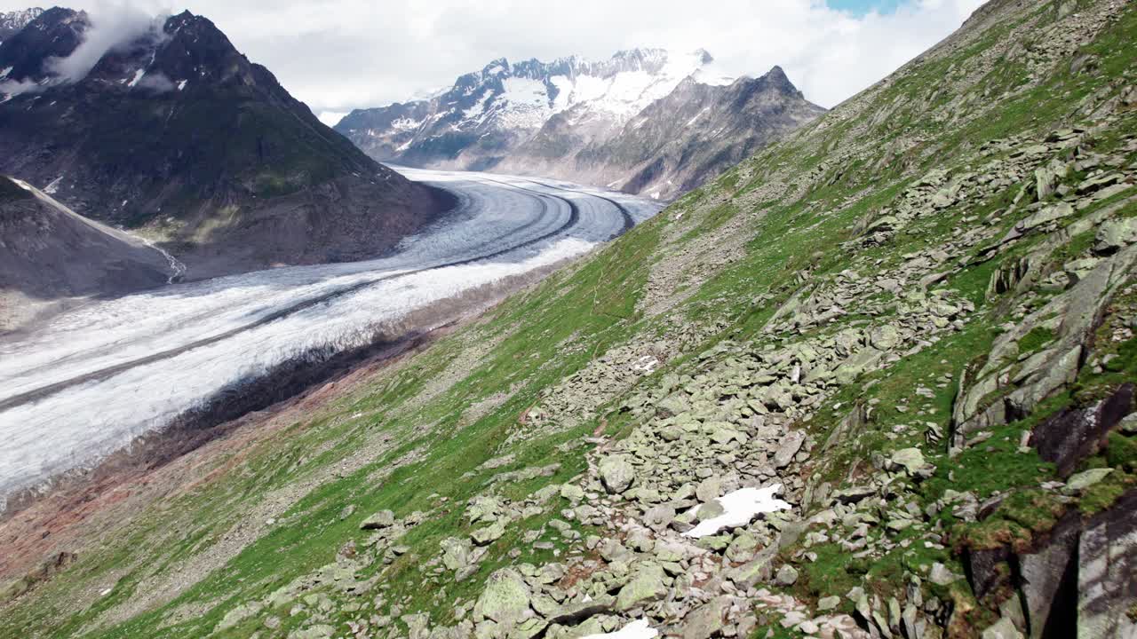 Cinematic aerial view of a lone hiker descending a mountain trail above a vast alpine glacier, emphasizing scale, exploration, and humanity’s presence in a dramatic high‑altitude landscape Switzerland