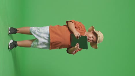 asian tourist boy with a hat using a tablet and smiling while standing on the green screen background. boy researcher examines something, travel tourism adventure concept, full body