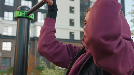 side view of gym goer working out on pull up bar in urban park environment, chest touching bar during intense exhaling to maintain balance and forearm strength while focusing on form and control