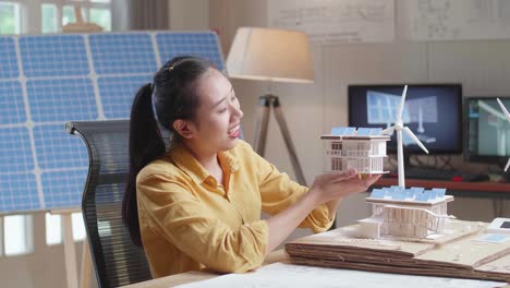 vista lateral de una mujer asiática sacudiendo positivamente la cabeza mientras sostiene y mira el modelo de una pequeña casa con panel solar
